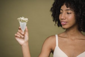 Happy young African American female model with curly hair in bra smiling and showing menstrual cup with gentle flowers against beige background