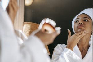 African American young female in white bathrobe applying moisturizing cream on face while standing in bathroom